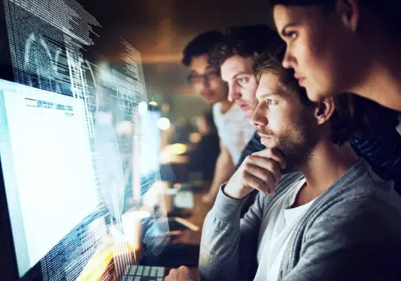 A group of four individuals closely reviewing code work on a number of computer screens, faces lit only by screens