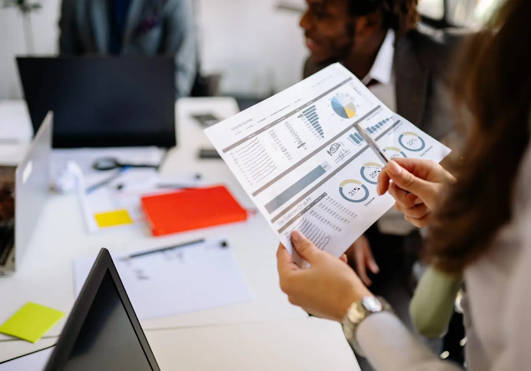 close up of paper with printed expense allocation numbers and graphics held by woman reviewing with pencil, other coworkers around table and paperwork blurred in the background