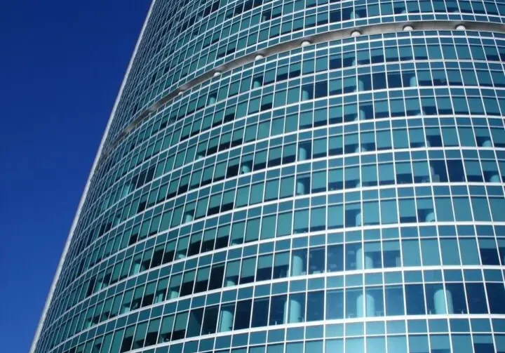 Tall glass office building from below with sun shining on the windows, deep blue sky behind.