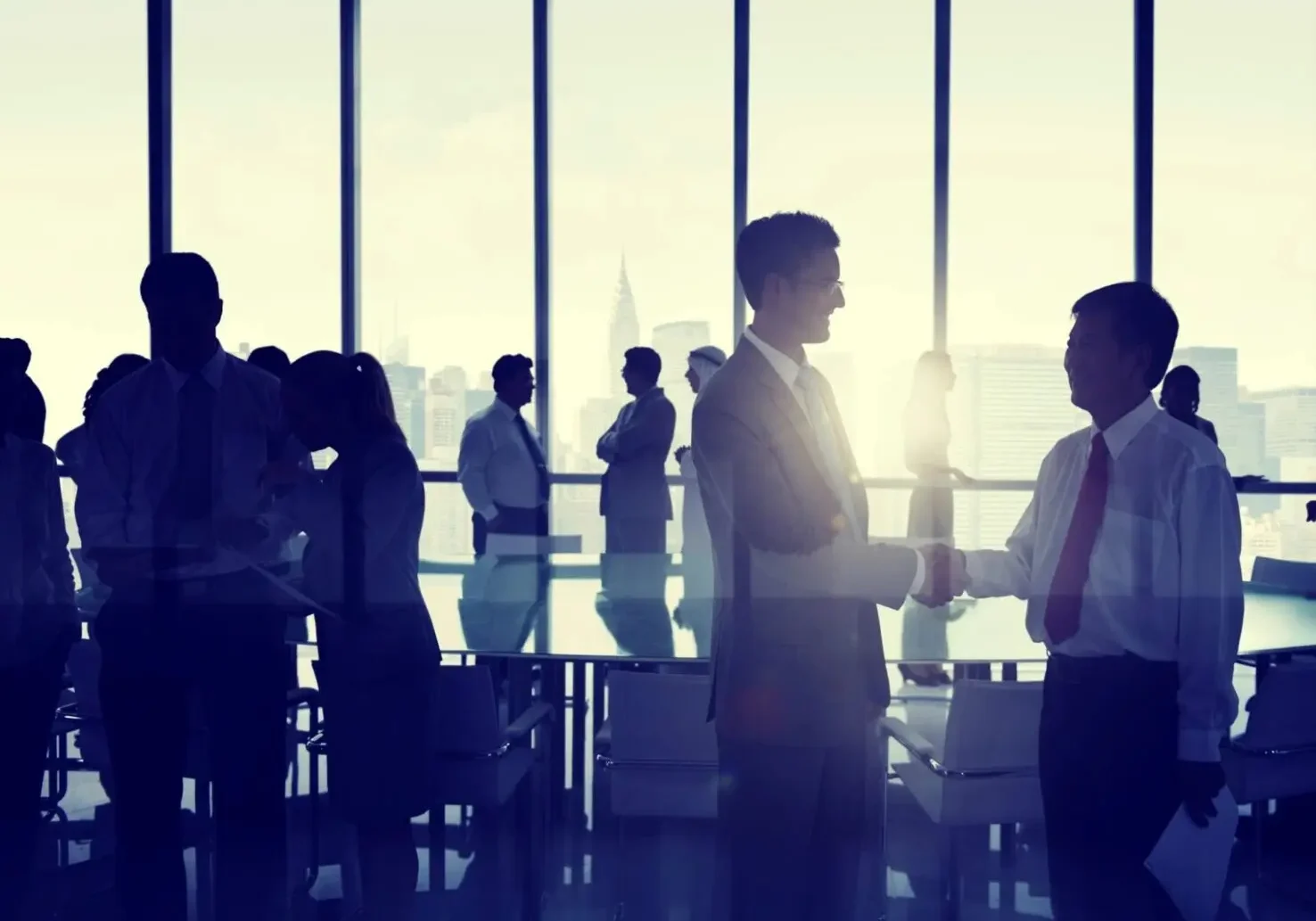 Silhouette of business men shaking hands in front of a table surrounded by other various business people talking in multiple groups with large windows spilling in light from the background