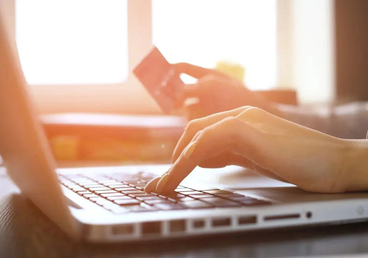 Close up view from left side of a laptop with a woman's hands typing with the left and holding a credit card with the right, light in the background